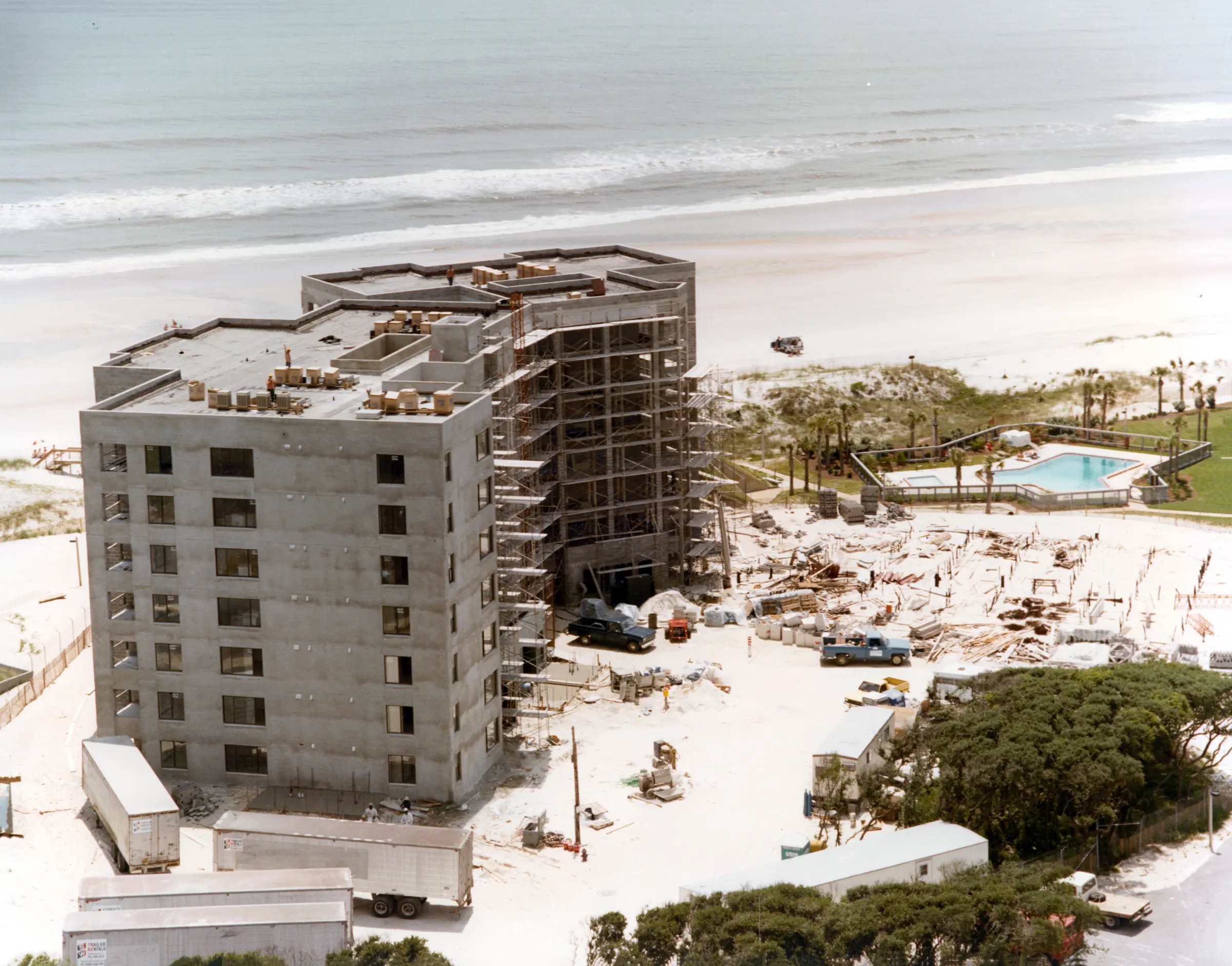 A concrete building under construction near the beach, with scaffolding, trucks and trailers around it and a finished pool beside it