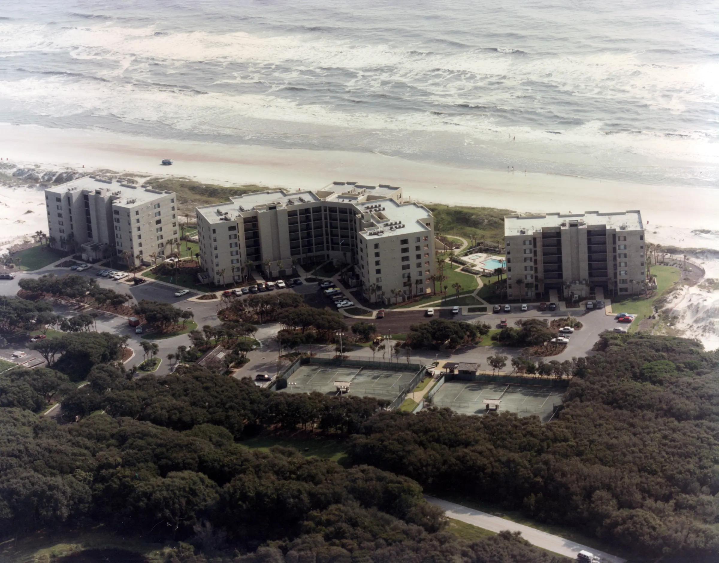 Aerial view from inland of the three completed buildings, with tennis courts and woods in the foreground and the surf beyond