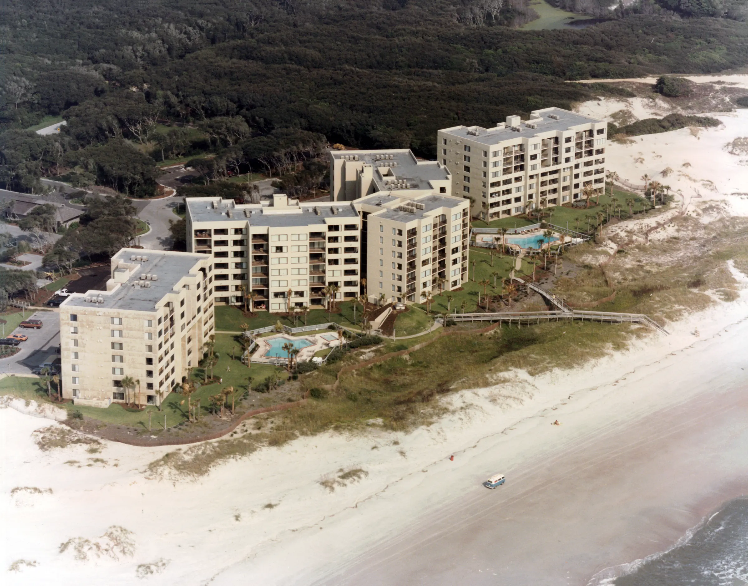 Aerial view of Amelia Surf & Racquet Club from over the ocean: three oceanfront buildings, two pools and a boardwalk over the dunes, dense woods behind, and a Volkswagen bus driving on the beach in the foreground