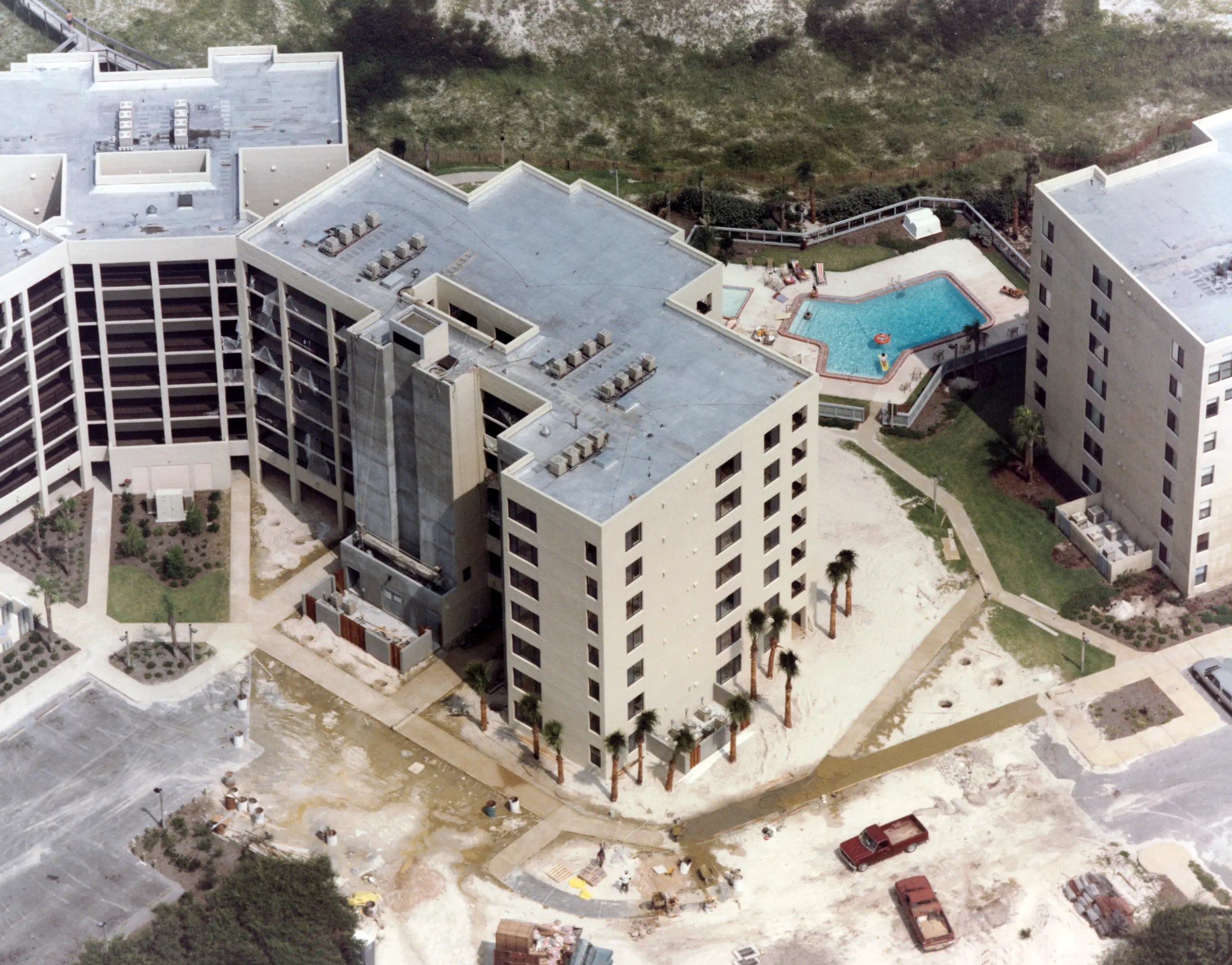 Aerial view of a newly completed building and its pool, with sandy, unfinished grounds and pickup trucks in the foreground