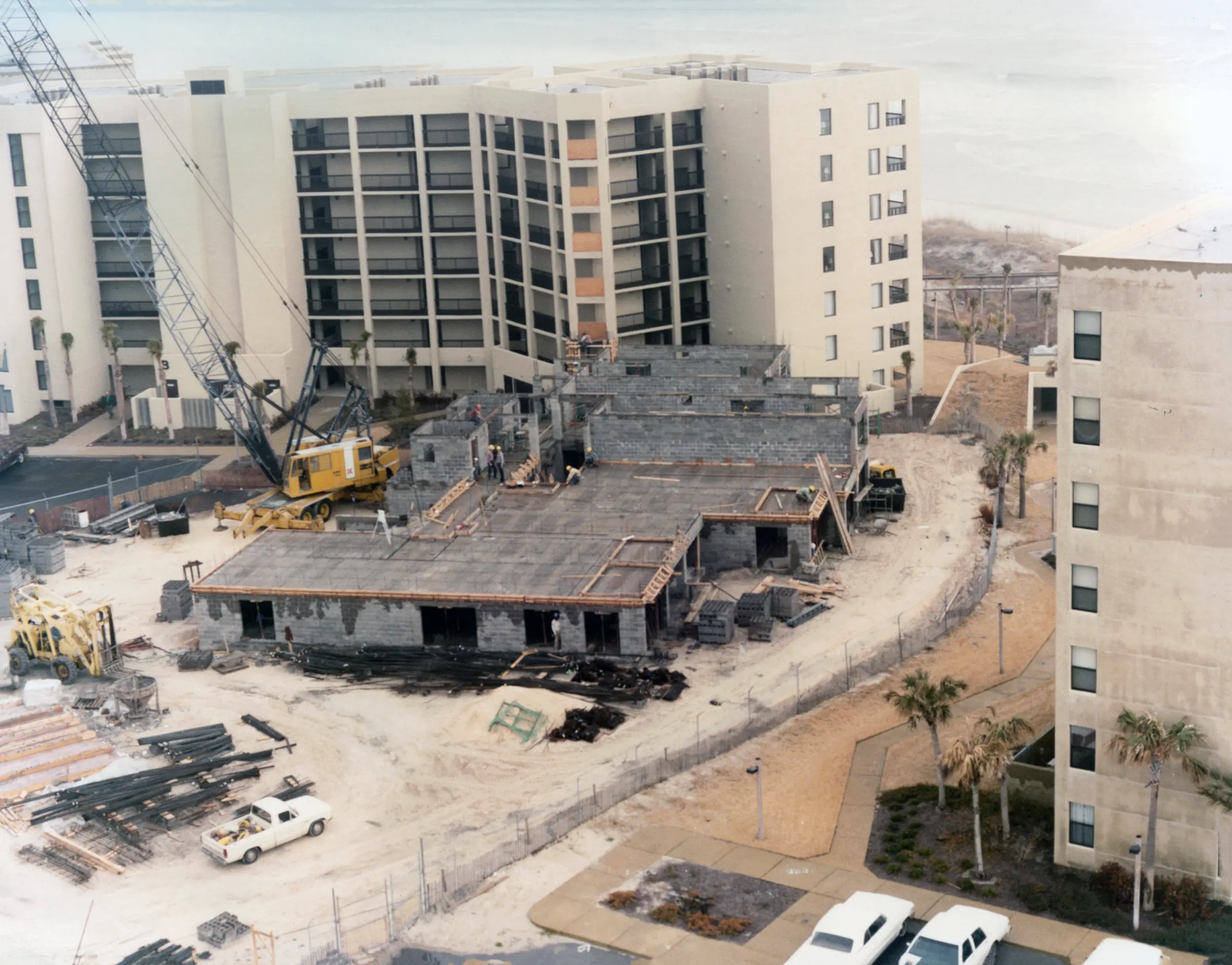 A crane building a low structure between completed buildings, with steel and materials stacked on the site