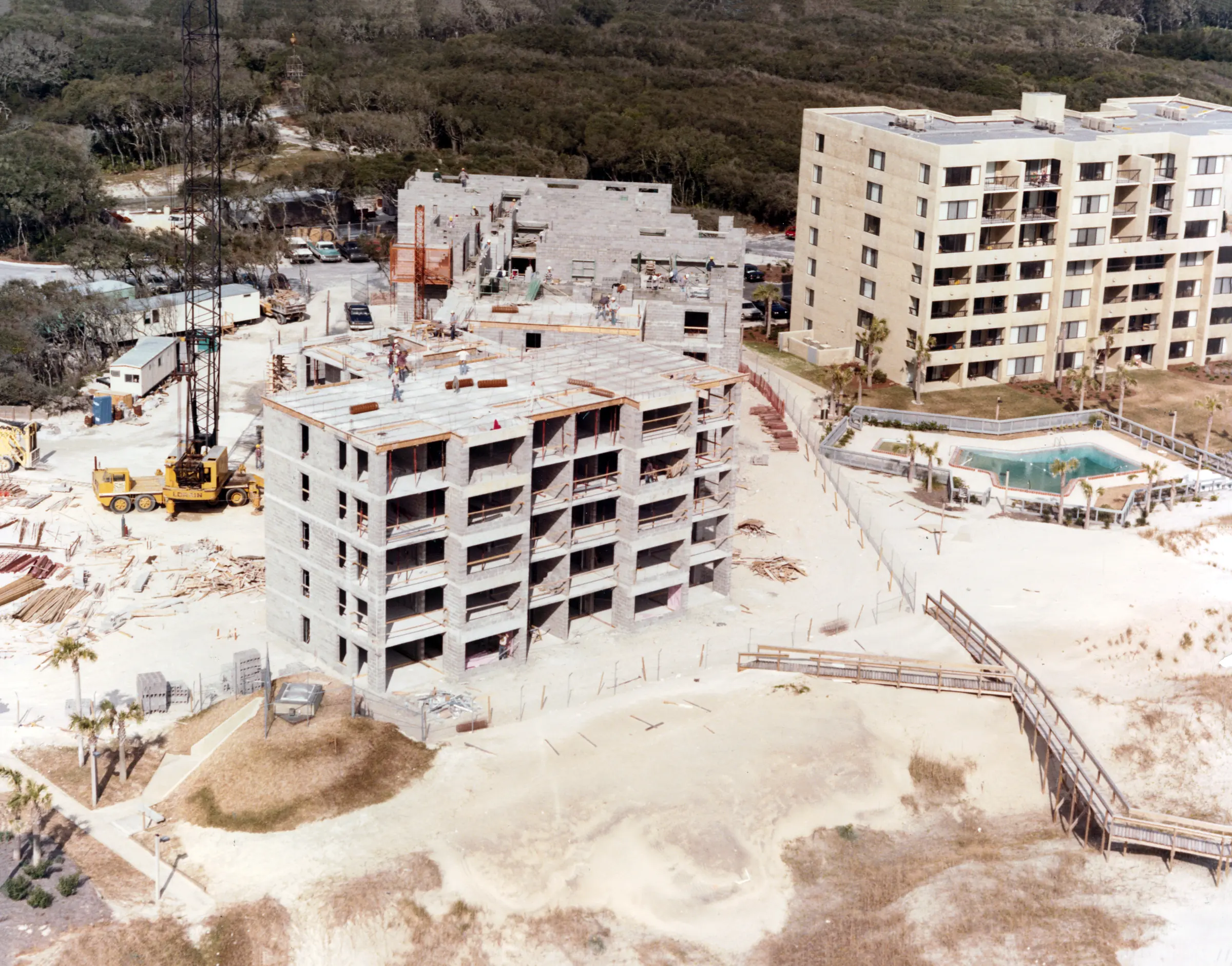 A building under construction with a crane beside it, its concrete floors partly framed, next to a completed building and pool