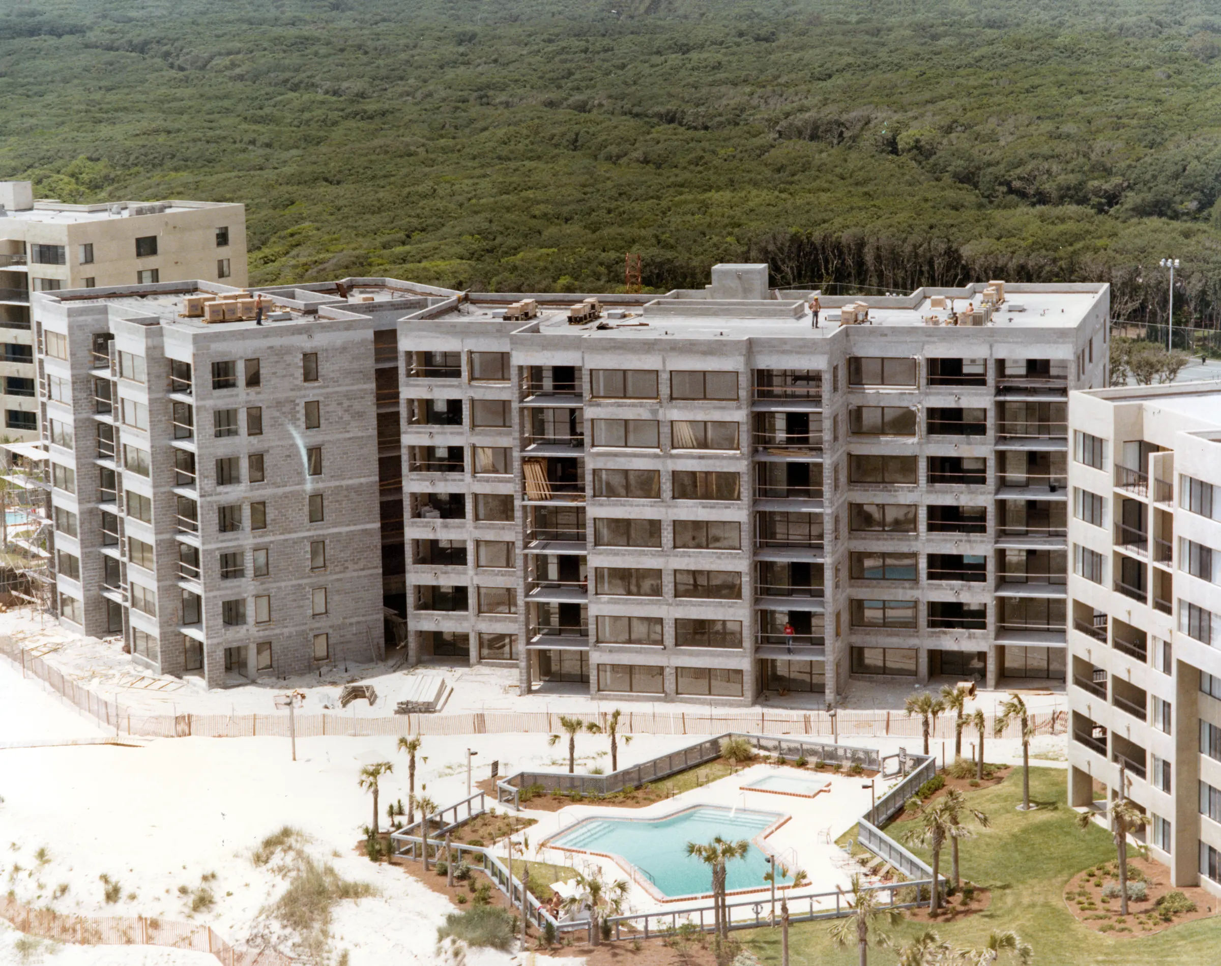 A building in the final stages of construction behind a landscaped pool, with dense woods stretching to the horizon