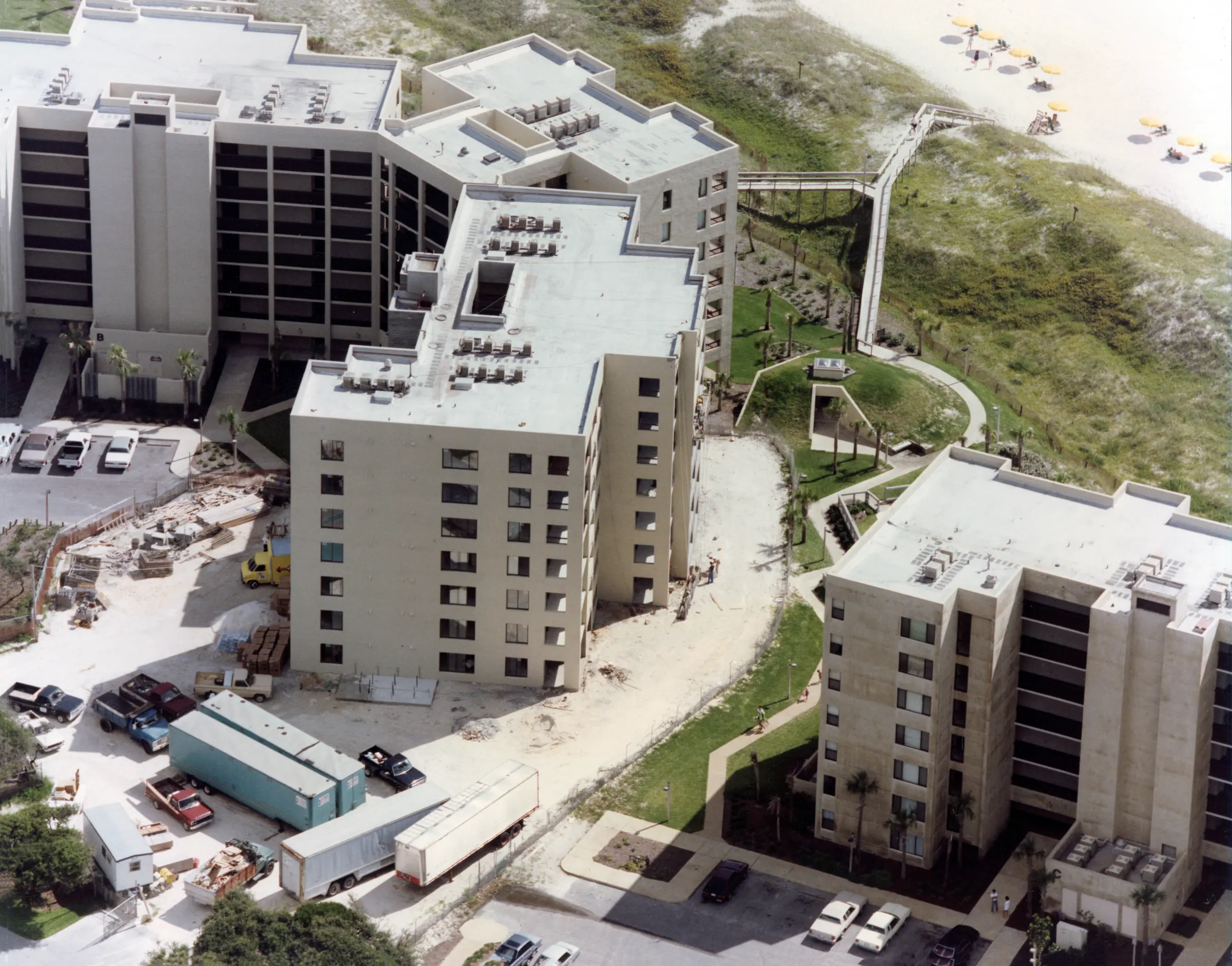 Aerial view of the completed buildings, landscaped grounds and a boardwalk over the dunes, with beach umbrellas lined up on the sand