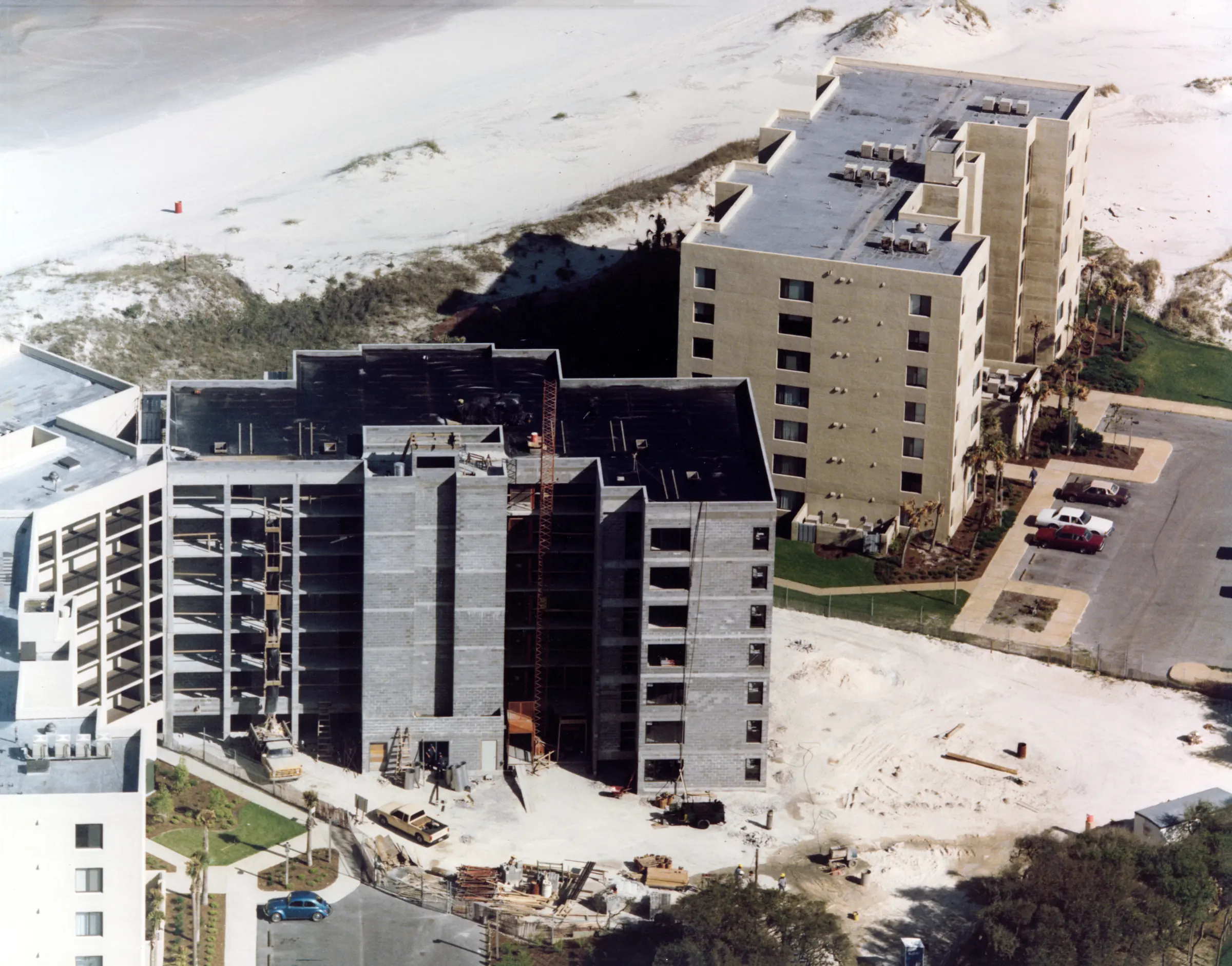 Aerial view of a building under construction beside a completed building, with dunes and the beach beyond