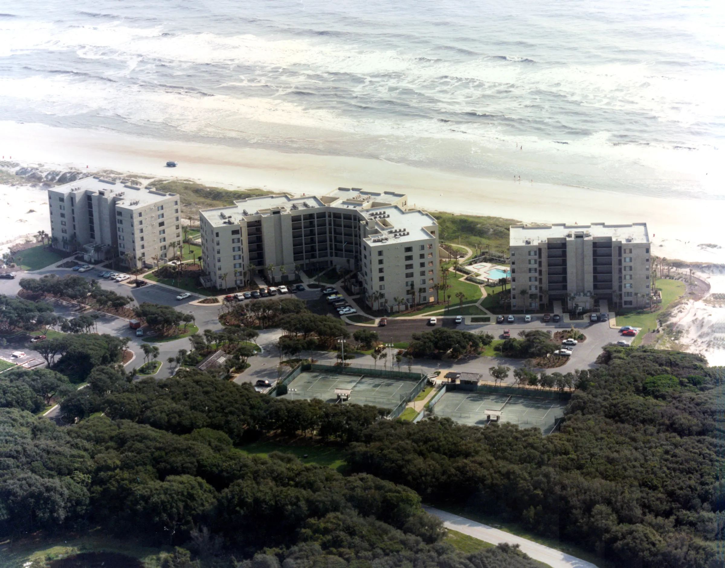 Aerial view from inland of the three completed buildings of Amelia Surf & Racquet Club, with tennis courts and woods in the foreground and the beach and surf beyond