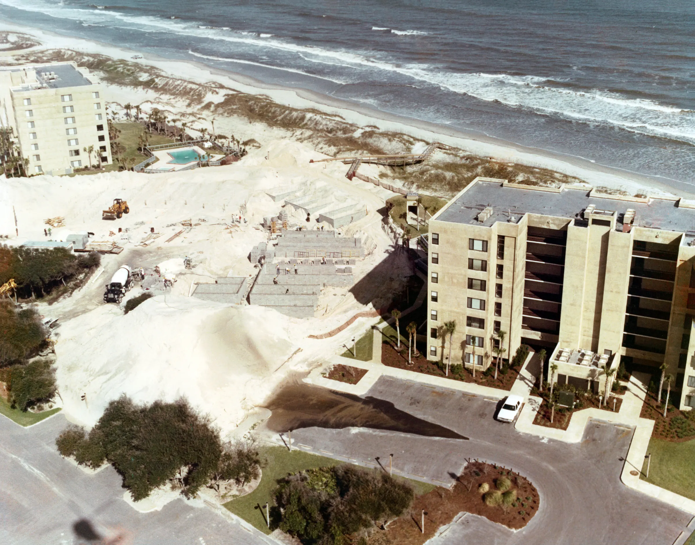 Aerial view of concrete foundation work in open sand beside a completed building, with a cement truck and a front loader on site