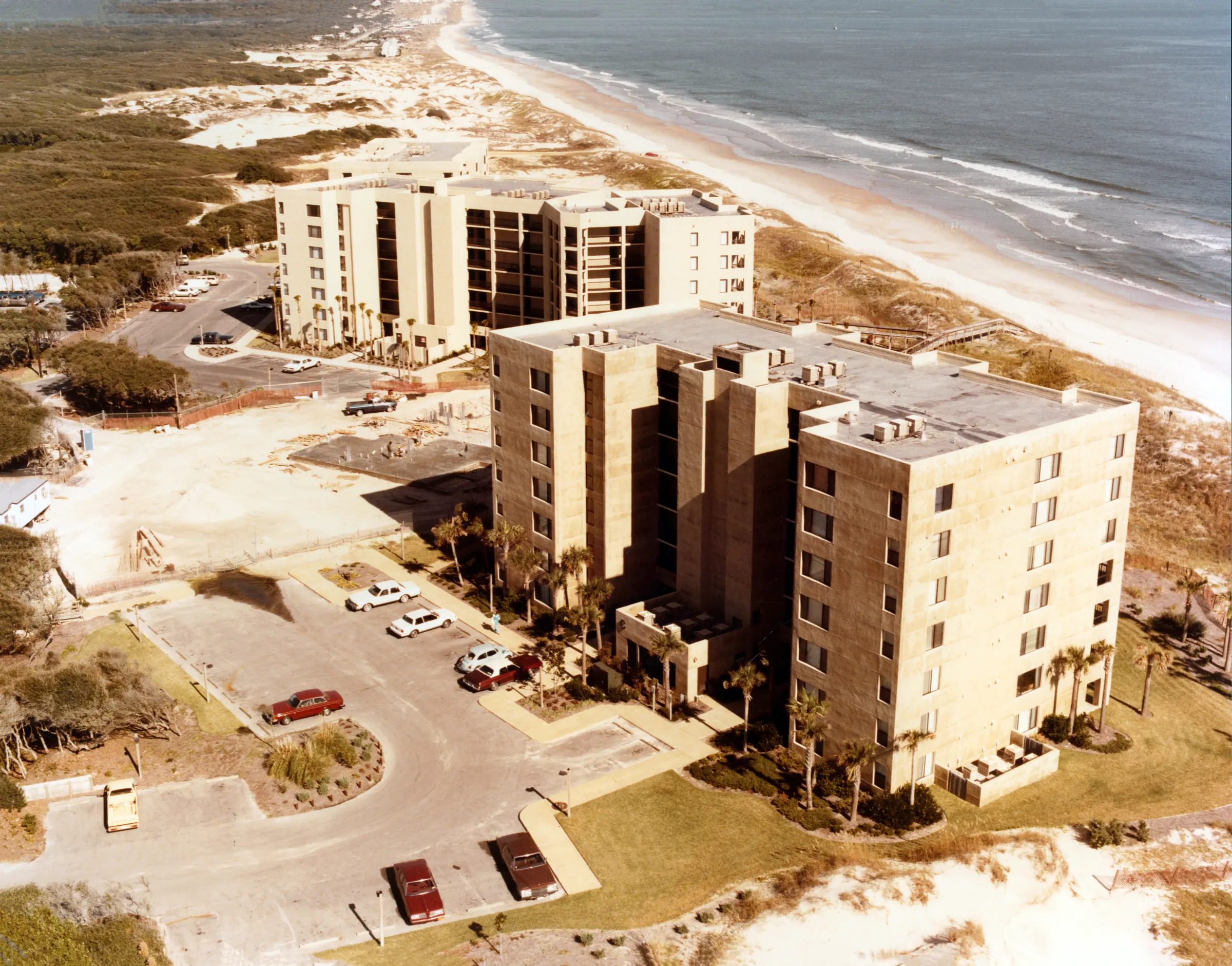 Aerial view of two completed Surf & Racquet buildings and their parking area, with the beach on the right and undeveloped dunes and woods stretching into the distance