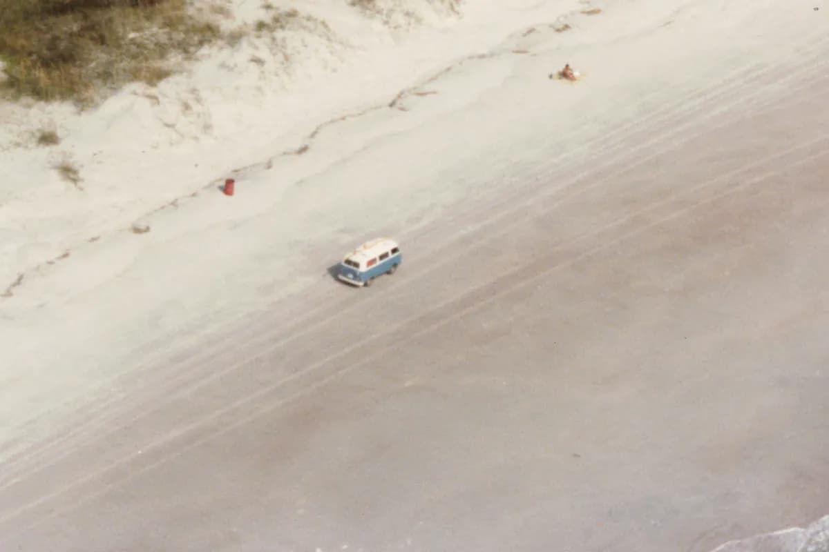 Close-up of a blue-and-white Volkswagen bus driving on the hard-packed sand below the dunes