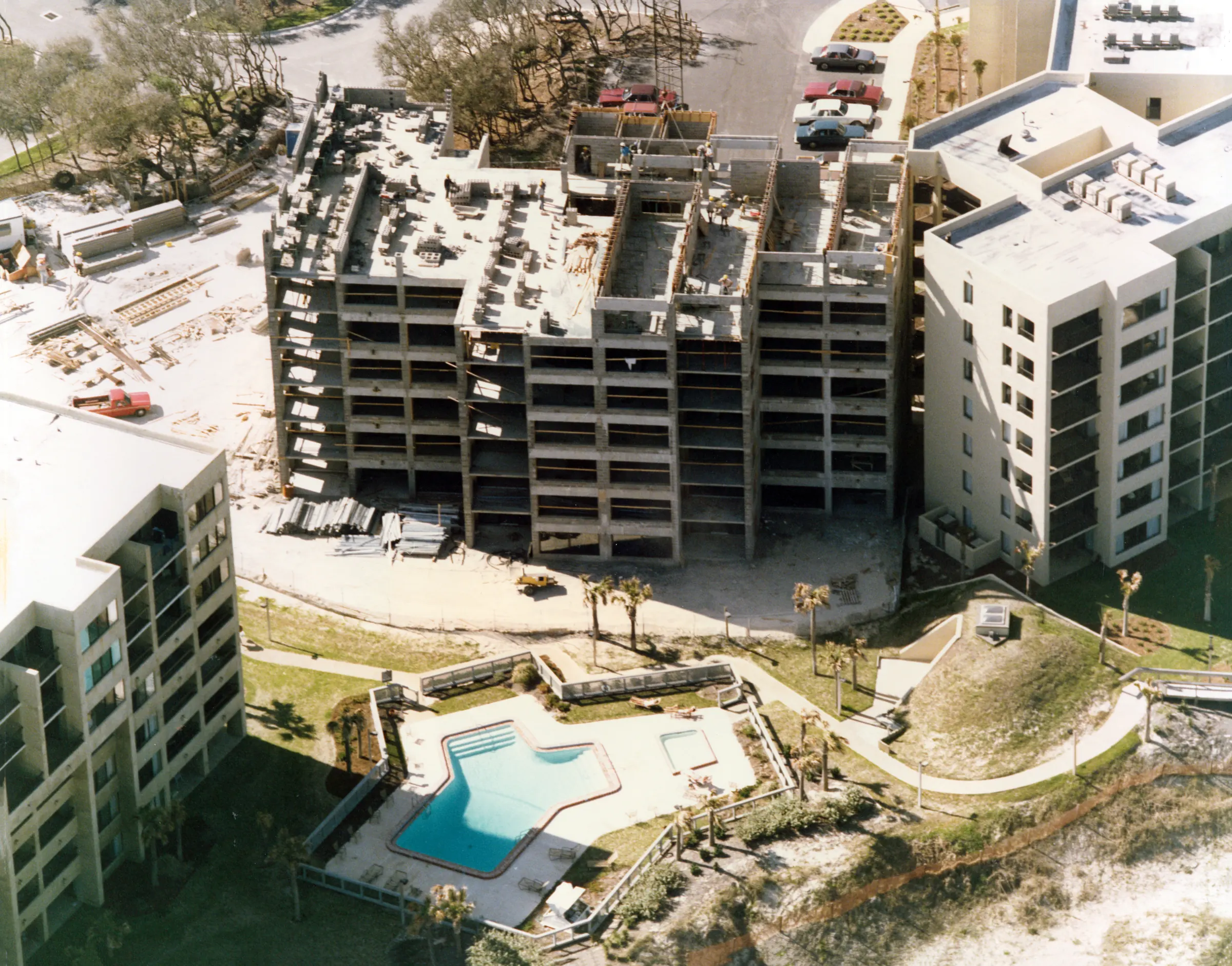 Aerial view of an unfinished concrete building frame between two completed buildings, with a finished pool and deck in the foreground