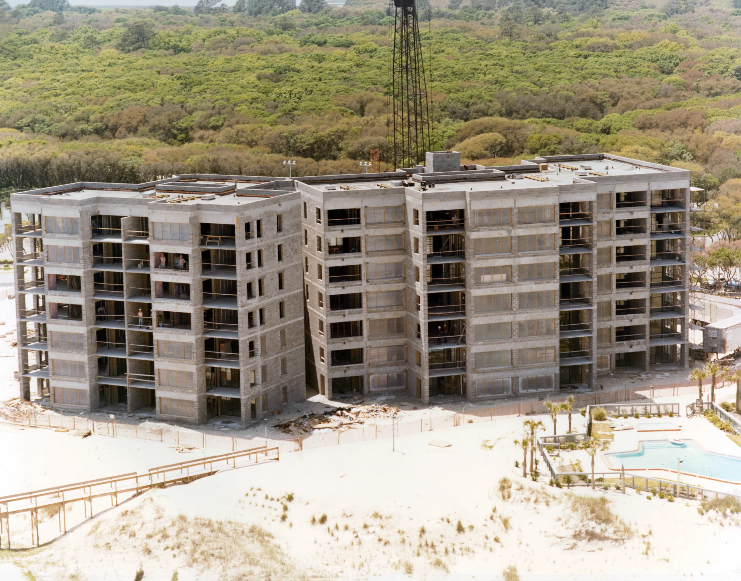 A seven-story concrete building under construction, seen from over the beach, with a crane behind it, a pool to the right and woods beyond