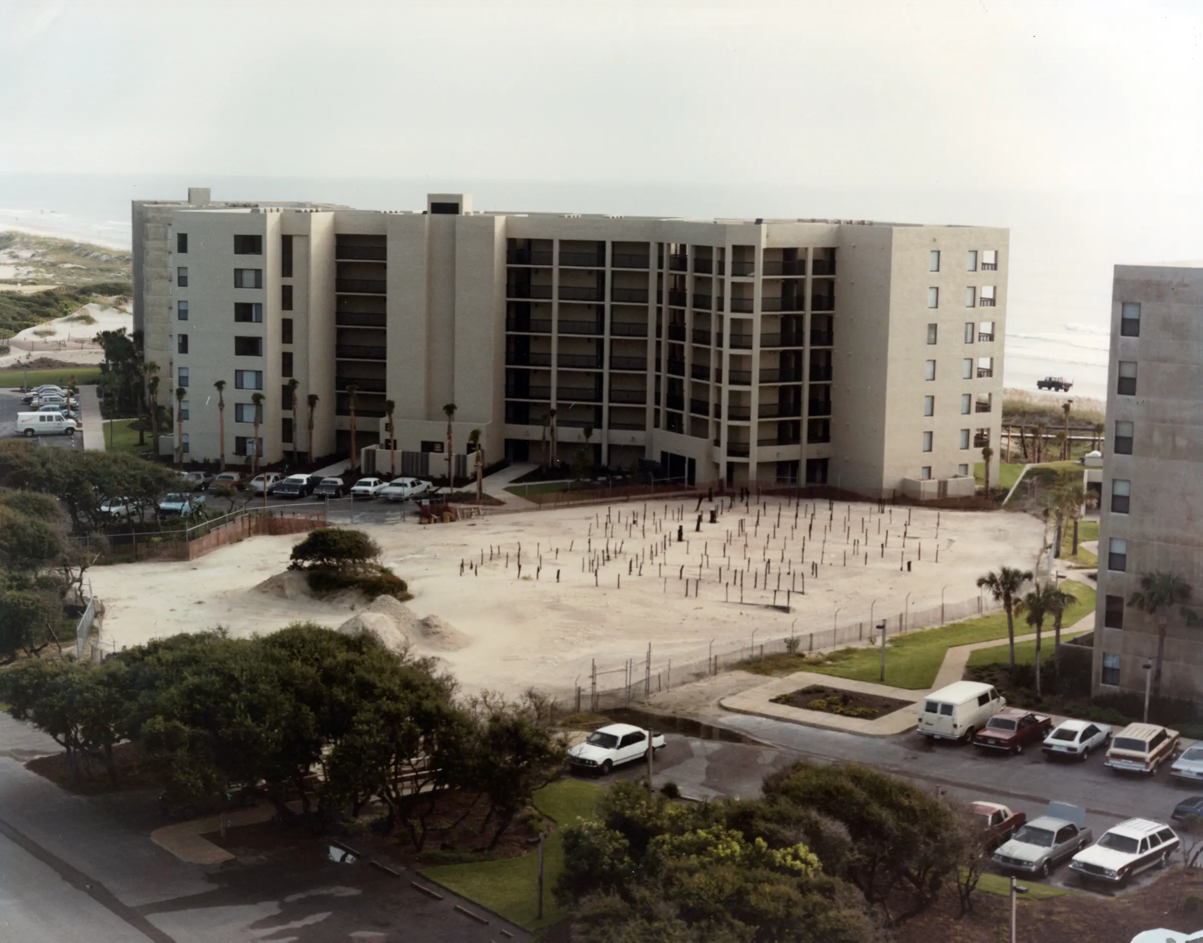 A completed Surf & Racquet building with cars in its parking lot, and a large cleared, sandy construction site in the foreground