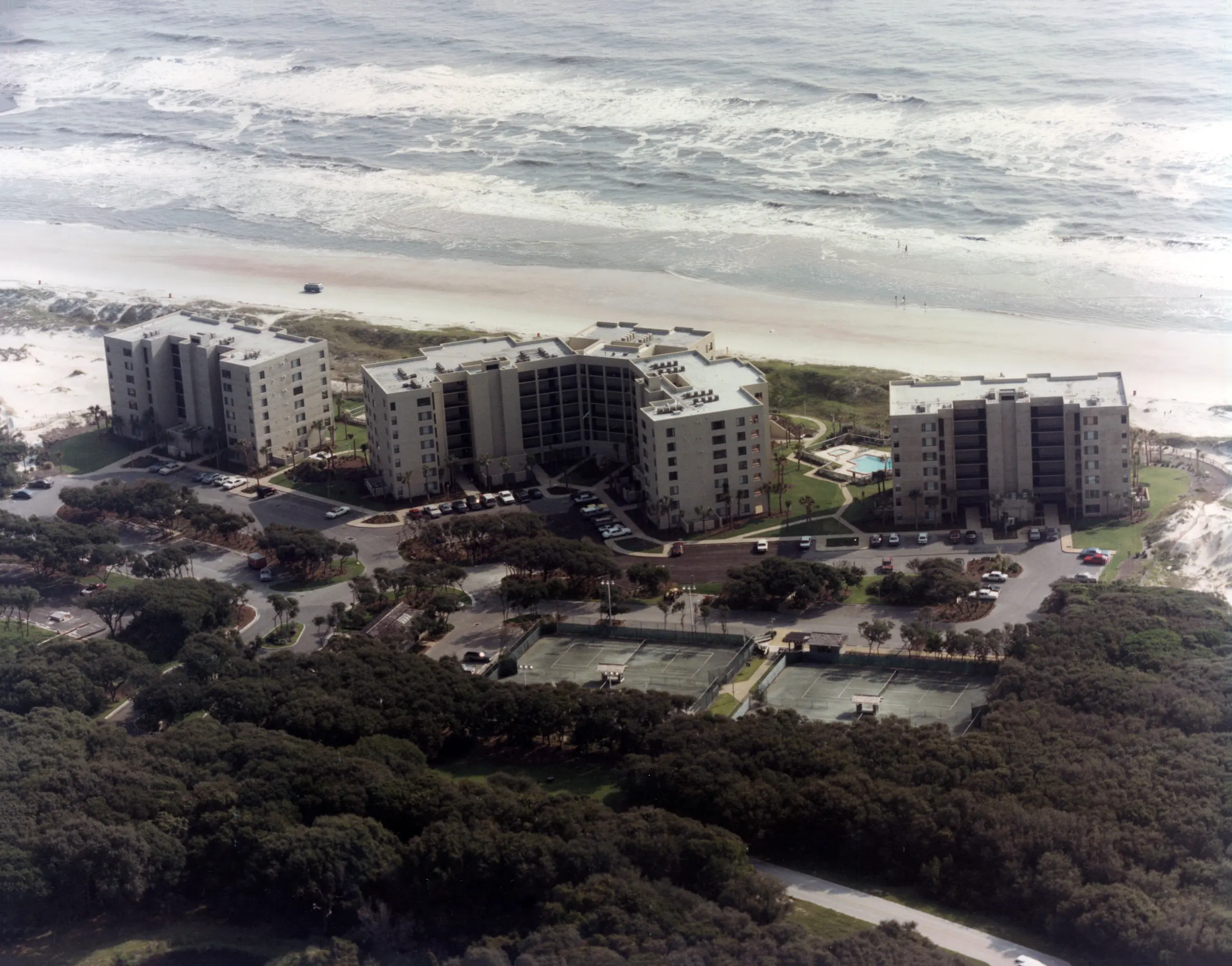 Wide aerial view of Amelia Surf & Racquet Club’s three buildings on the oceanfront, surrounded by dense woods, with tennis courts in the foreground and the Atlantic surf beyond