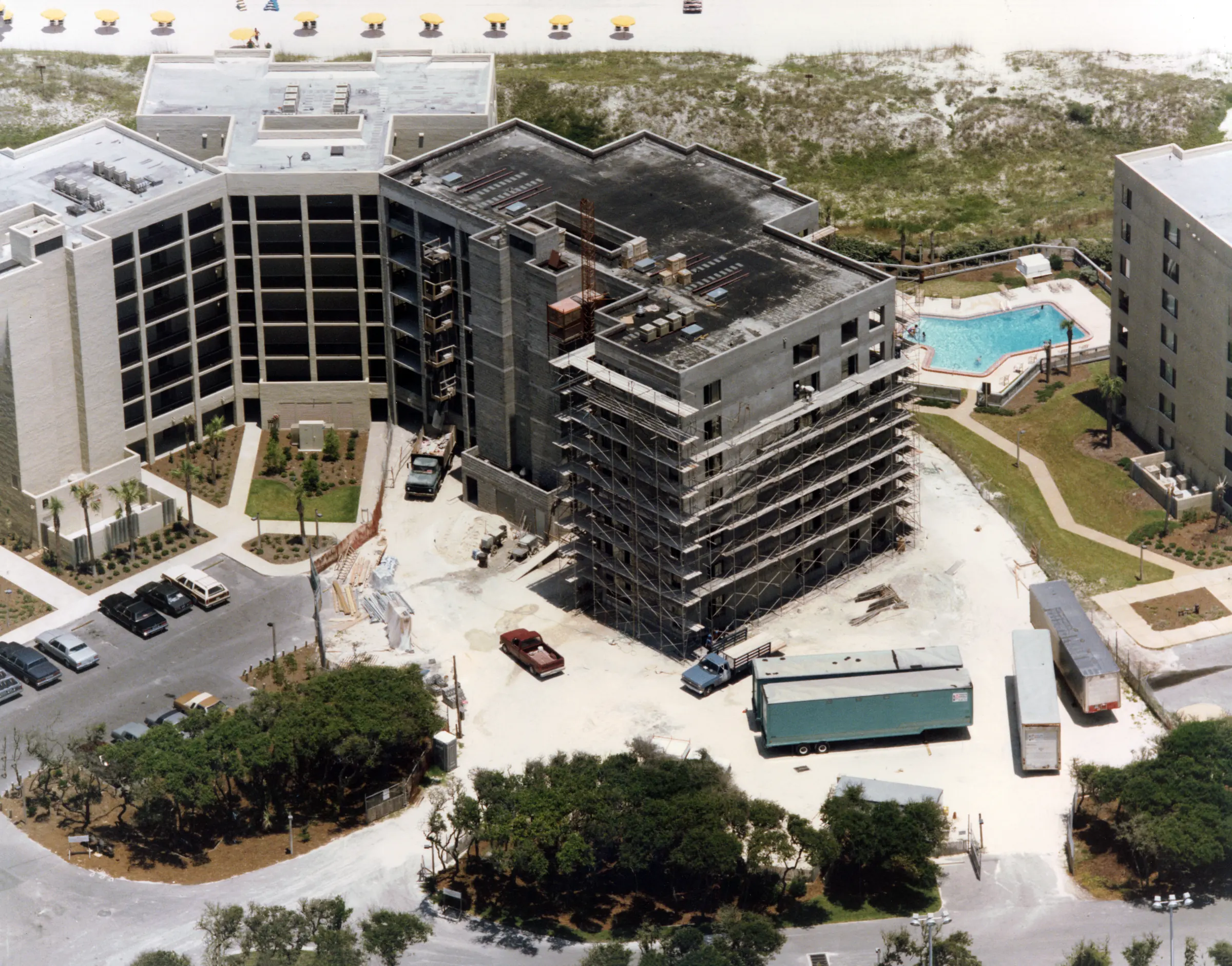Aerial view of a building still in scaffolding, surrounded by completed buildings, a pool, parking and construction trailers